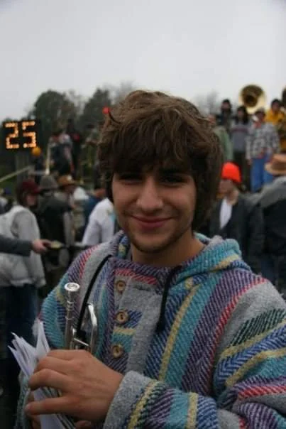 A young Christopher Jager in a striped hooded sweater holding a trumpet and sheet music at an outdoor football game, a crowd and scoreboard behind him